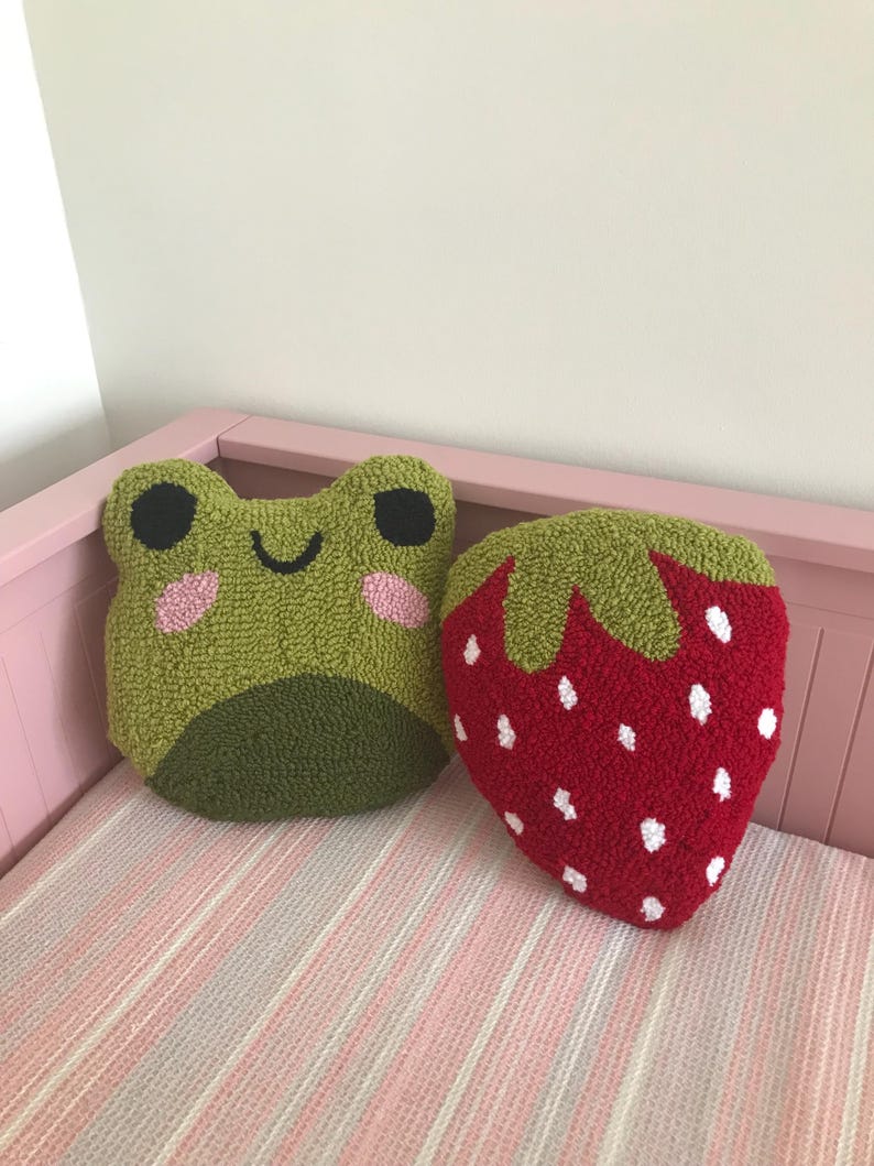 Puede incluir: Dos almohadas de felpa, una con forma de rana verde con una cara sonriente y la otra con forma de fresa roja con semillas blancas.