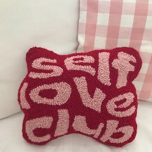 May include: A red, textured cushion with the words "SELF LOVE CLUB" in light pink. The cushion is rectangular with rounded edges. The background includes a white pillow and a pink and white checkered pillow.