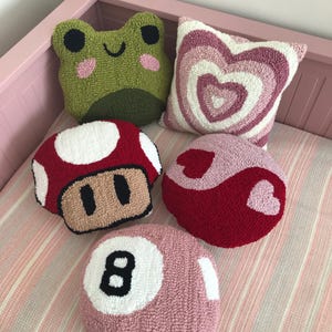 Puede incluir: Cinco almohadas decorativas de colores y cosidas a mano con diferentes dise&ntilde;os. Las almohadas est&aacute;n hechas de una tela suave y mullida y presentan una rana verde, un hongo rojo, un coraz&oacute;n rosa, un s&iacute;mbolo yin-yang rojo y rosa y una bola de billar rosa.