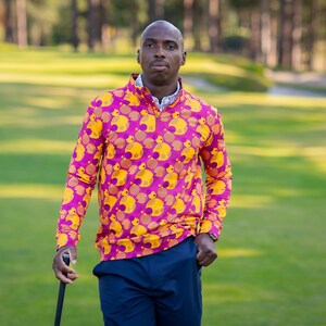 Peut inclure: Une chemise de golf à manches longues avec un motif de canards jaunes sur fond magenta. La chemise a un col zippé et est portée avec un pantalon bleu marine. La personne tient un club de golf sur un parcours vert.