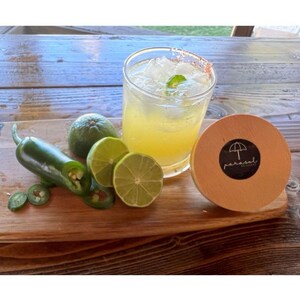 May include: A cocktail in a glass with ice and a mint garnish, with a salt-rimmed edge. Limes, a whole jalape&ntilde;o, and sliced jalape&ntilde;o peppers are on a wooden cutting board. A wooden coaster with a logo is on the right.
