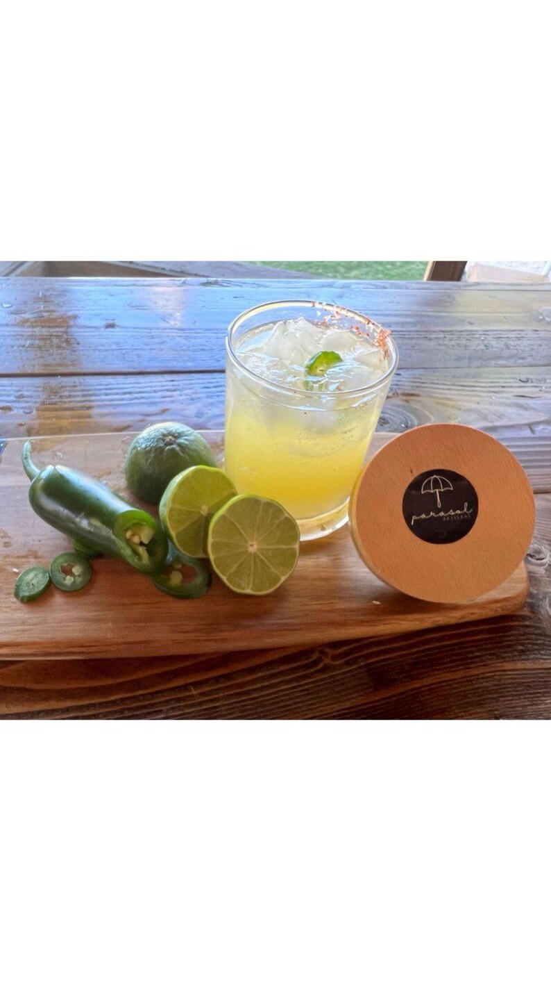 May include: A cocktail in a glass with ice and a mint garnish, with a salt-rimmed edge. Limes, a whole jalape&ntilde;o, and sliced jalape&ntilde;o peppers are on a wooden cutting board. A wooden coaster with a logo is on the right.