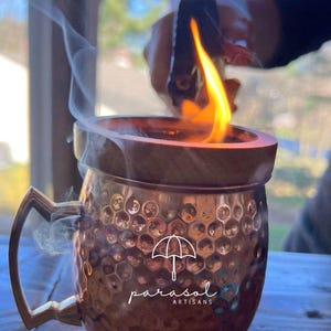 May include: A copper mug with a wooden lid and handle, featuring a hammered texture. The mug has the text "parasol ARTISANS" on the front. Flames are visible above the mug, with smoke rising. The mug is on a wooden surface.