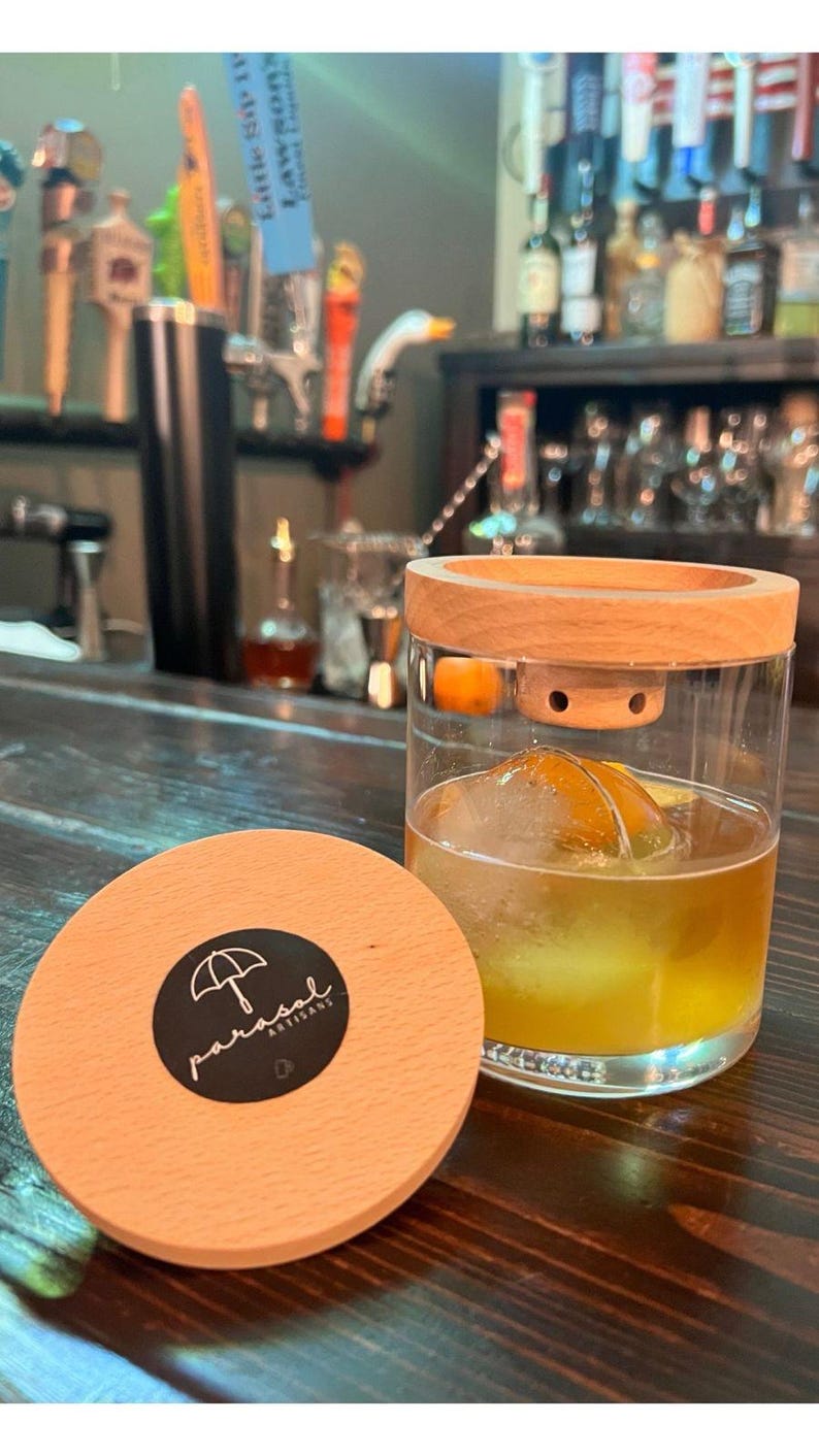 May include: A cocktail glass with a wooden lid and a large ice sphere containing an orange slice. A wooden coaster with the text "parasol artisans" and an umbrella logo is in the foreground. The drink is amber-coloured.