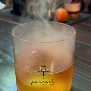 May include: A cocktail in a clear glass with a smoky vapor rising from it. The drink is amber-coloured, and the glass has the "parasol artisans" logo. An orange and a bottle of liquor are in the background.