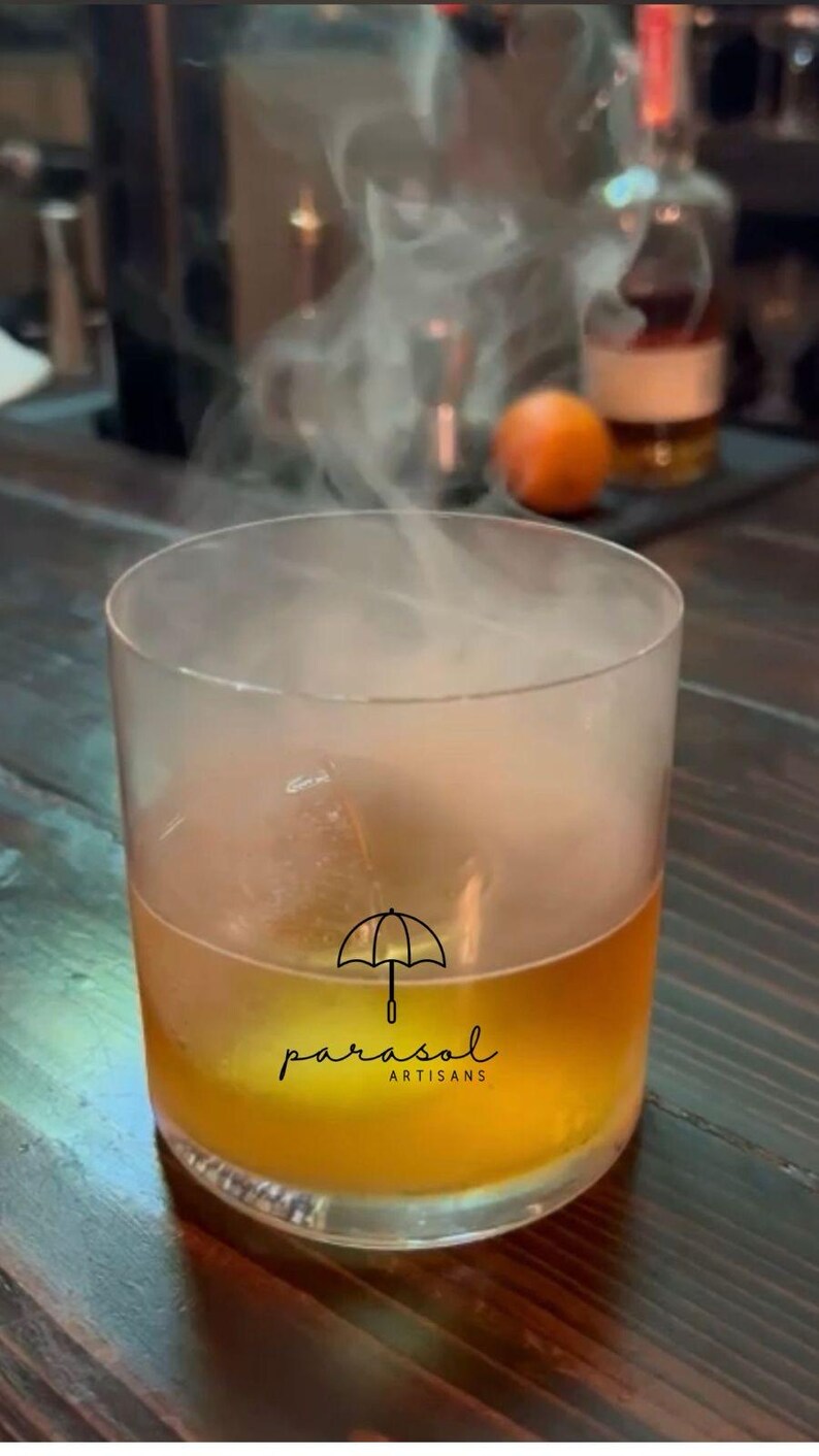 May include: A cocktail in a clear glass with a smoky vapor rising from it. The drink is amber-coloured, and the glass has the "parasol artisans" logo. An orange and a bottle of liquor are in the background.