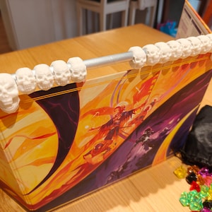 May include: A fantasy game accessory with a vibrant illustration. The top edge is decorated with a row of white skull-shaped ornaments. A black bag and colourful gems are also visible.
