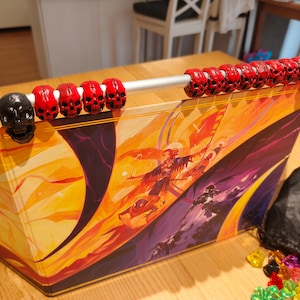 May include: A fantasy role-playing game box featuring a vibrant illustration. The box is decorated with red skull beads and a black skull. A black bag and colourful gems are also visible.