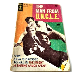 Könnte beinhalten: Vintage Taschenbuchcover für "The Man From U.N.C.L.E." mit zwei Männern in Anzügen, einer hält eine Waffe. Das Cover hat ein rosa, grünes und orangefarbenes Farbschema mit dem Titel in großen, fetten Buchstaben. Text enthält "SIR" und "ILLYA IS DRESSED TO KILL IN THE KNIGHT IN SHINING ARMOR AFFAIR."