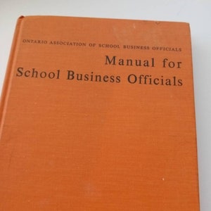 May include: An orange book titled "Manual for School Business Officials" by the Ontario Association of School Business Officials. The text is in black, and the book has a textured cover.
