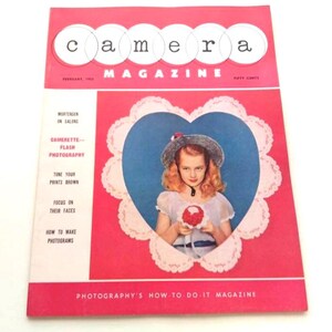 Puede incluir: Portada de la revista "Camera" de febrero de 1953. La portada presenta a una niña con sombrero dentro de un marco en forma de corazón. El título de la revista está en letras blancas sobre un fondo rojo. La revista es una guía de fotografía.