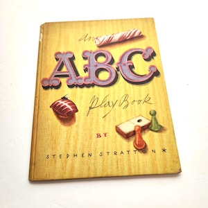 May include: A vintage ABC Play Book by Stephen Stratton. The cover features the title "ABC" in large, purple letters with a red outline. Illustrations include a candy cane, a Christmas ornament, and game pieces.