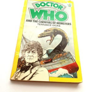 May include: A vintage paperback book titled "Doctor Who and the Carnival of Monsters" by Terrance Dicks. The cover features a green and white logo, an illustration of a monster, a ship, and a portrait of a man in a suit.