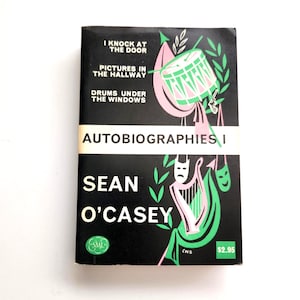 May include: A black book cover titled Autobiographies I by Sean O'Casey. The cover features white text, a green and pink illustration of a drum, a harp, and theatrical masks. The text reads I Knock at the Door, Pictures in the Hallway, and Drums Under the Windows.