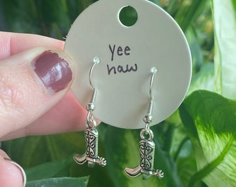 May include: A pair of silver cowboy boot earrings with a spur detail. The earrings are on a white card with the text "Yee Haw" written on it.