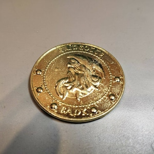 May include: A gold-colored coin with a raised image of a mythical creature with wings and a crown. The coin has the words "GRIFFITH" and "GADKO" inscribed around the edge.