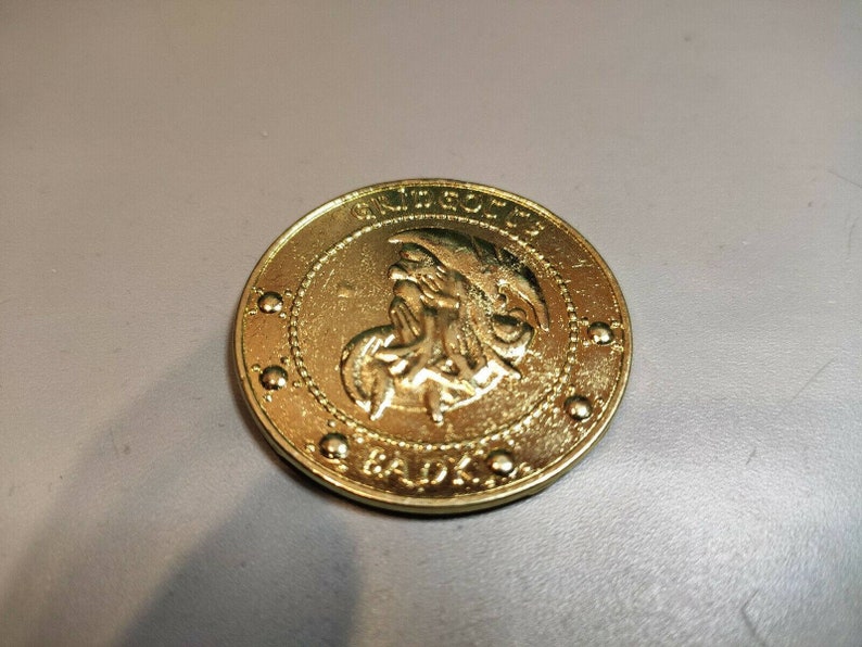 May include: A gold-colored coin with a raised image of a mythical creature with wings and a crown. The coin has the words "GRIFFITH" and "GADKO" inscribed around the edge.