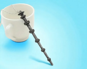 May include: A silver necklace with a detailed wand pendant. The wand is a dark silver color and has a textured surface. The necklace is laying on a white cup.