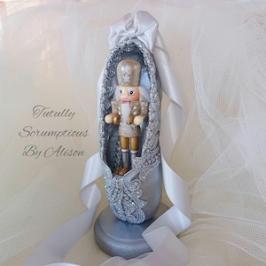 May include: A decorative silver ballet shoe, featuring a miniature nutcracker figure. The shoe is adorned with glitter and lace detailing, and has a white ribbon. The text "Tutully Scrumptious By Alison" is visible on the side.