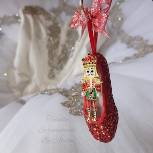May include: A red glitter ballet slipper ornament with a gold and jewel-toned nutcracker figure. The ornament has a red ribbon and bow at the top. The background is a white tutu.