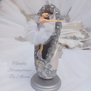 May include: A white porcelain ballerina figurine is posed in a dance position inside a silver and white decorated ballet shoe. The shoe is on a silver stand. The text "Tutully Scrumptious By Alison" is visible in the image.