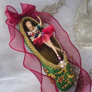 May include: A green satin ballet slipper with gold trim and rhinestones. A miniature ballerina doll is inside the slipper, wearing a red tutu and holding a miniature nutcracker doll.