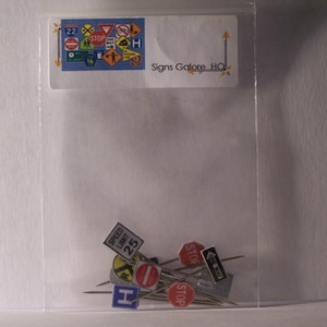 May include: A clear plastic bag containing miniature road sign pins. The pins feature various traffic symbols, including stop signs, speed limits, and directional arrows. The bag has a label that reads "Signs Galore HQ".
