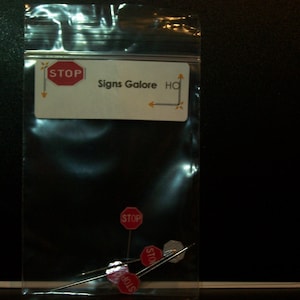 May include: A clear plastic bag containing a set of red and white "STOP" signs on small metal pins. The bag has a label that reads "Signs Galore HQ".