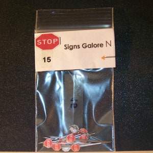 May include: A clear plastic bag containing 15 small red and white "STOP" signs with silver pins. The bag has a label that reads "Signs Galore N" and "15".