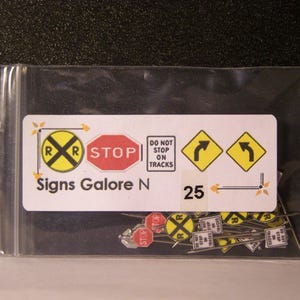 May include: A clear plastic bag containing a set of 25 miniature road signs. The signs include a railroad crossing sign, a stop sign, a do not stop on tracks sign, and two right turn signs. The bag is labeled "Signs Galore N 25".
