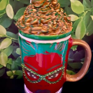 Puede incluir: Una taza de cerámica roja con un diseño verde y dorado. La taza presenta una cara estilizada con gafas de purpurina verde. La taza tiene un asa dorada y una tapa verde y dorada.