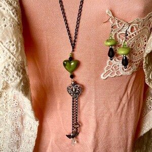 May include: A long green and black necklace with a heart-shaped pendant and a key charm. The necklace is made of metal and beads. The pendant is green and the key charm is silver.