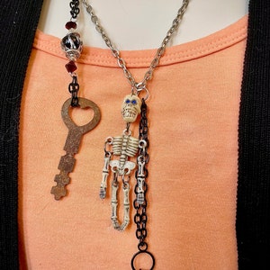 Peut inclure: Un collier en chaîne argentée avec un pendentif squelette, un pendentif clé et un pendentif à perles aux accents rouge et noir.