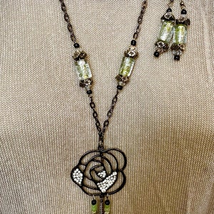 May include: A long, brown chain necklace with a rose-shaped pendant. The pendant is made of metal and has a filigree design. The necklace is accented with green and black beads. Matching earrings with green and black beads are also included.