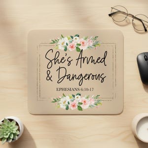 Peut inclure: Tapis de souris rectangulaire beige avec le texte "She's Armed & Dangerous" et "Éphésiens 6:10-17" en écriture noire. Le tapis est orné de compositions florales de roses roses et blanches. Une souris noire et des lunettes sont visibles.
