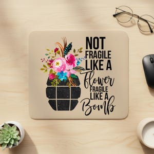 May include: A beige mousepad with a floral design and the text "NOT FRAGILE LIKE A FLOWER FRAGILE LIKE A BOMB." The floral design includes pink, blue, and red flowers, with a black grenade shape. A black computer mouse and glasses are visible.