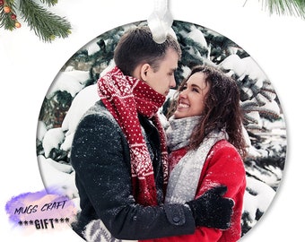 Peut inclure: Une boule de Noël ronde et blanche personnalisée représentant un couple dans un paysage d'hiver enneigé. Le couple porte des écharpes rouges et blanches et se regarde. La boule est idéale pour un cadeau de Noël romantique.