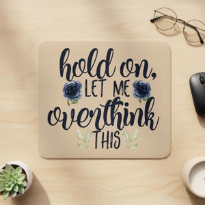 May include: A beige mousepad with the text "hold on, let me overthink this" in dark blue script. The design includes dark blue rose illustrations and green leaf accents. A computer mouse and glasses are visible.