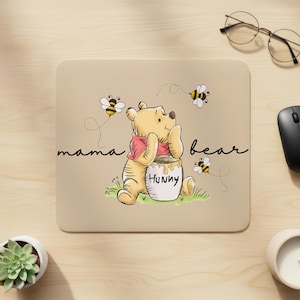 May include: Beige mousepad featuring a cartoon illustration of Winnie the Pooh with a honey pot. The text "mama bear" is written on the left side, and "bear" on the right. Bees are flying around the bear. A computer mouse and glasses are visible.