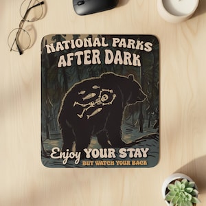 May include: A rectangular mousepad with a dark forest scene and a black bear silhouette. The bear has a skeleton design on its back. Text reads "NATIONAL PARKS AFTER DARK" and "ENJOY YOUR STAY BUT WATCH YOUR BACK."