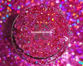Könnte beinhalten: Ein Glasbehälter gefüllt mit leuchtend pinkfarbenem Glitzer. Der Glitzer hat verschiedene Formen und Größen und reflektiert das Licht mit einem Regenbogeneffekt. Der Hintergrund ist ein verschwommener Bereich aus ähnlichem Glitzer, der einen funkelnden, festlichen Look erzeugt. Der Text "ISLAND GLITTER" ist sichtbar.