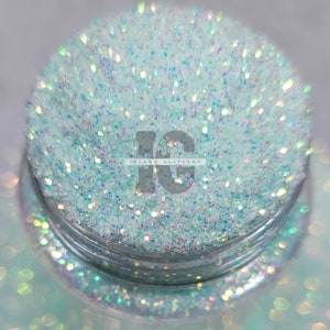 May include: A jar filled with iridescent glitter. The glitter is a mix of light blue and clear, with a sparkling, reflective quality. The text "ISLAND GLITTERS" is visible on the jar.