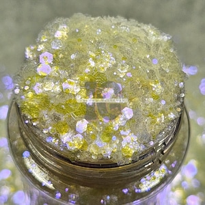 May include: A close-up of a glass jar filled with iridescent yellow glitter. The glitter is a mix of small and large hexagons, and it has a subtle shimmer.