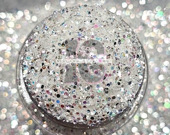 Può includere: Primo piano di un barattolo trasparente pieno di glitter iridescente. Il glitter è un mix di colori argento, bianco e arcobaleno. Il barattolo è appoggiato su un letto di glitter sciolto.