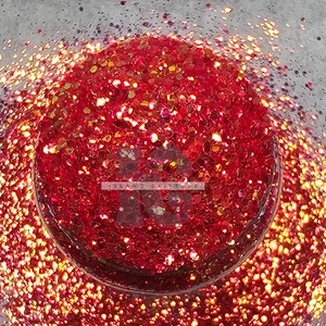 May include: A close-up of red glitter with a gold iridescent sheen. The glitter is in a pile and appears to be in a bowl or container.