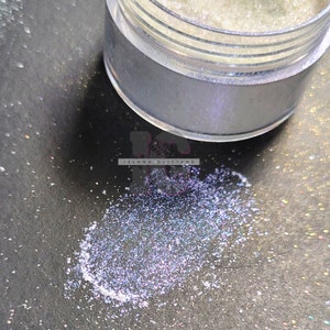 Op de afbeelding: Een klein, doorzichtig potje gevuld met iriserende zilveren glitter. De glitter valt uit het potje op een donker oppervlak.