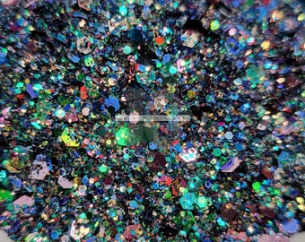 May include: A close-up of a mixture of iridescent glitter in various shades of blue, green, and purple. The glitter is scattered on a white surface.