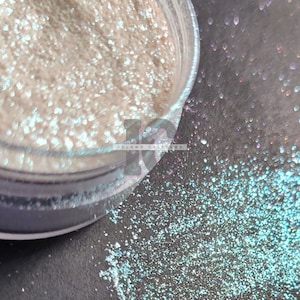 May include: A close-up of a jar of iridescent glitter. The glitter is a light blue color with a white iridescent sheen. The glitter is spilling out of the jar onto a black surface.