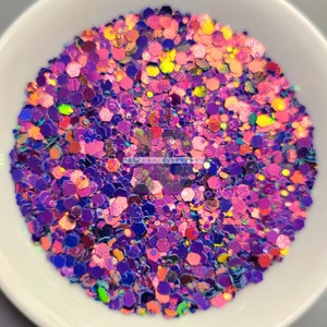 May include: A close-up of a bowl filled with iridescent glitter in shades of pink, purple, and blue. The glitter is in various shapes and sizes, creating a shimmering effect.