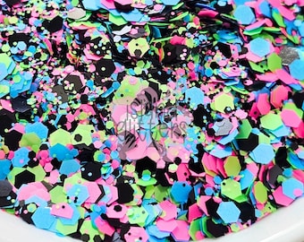 Könnte beinhalten: Nahaufnahme eines Haufens bunter Glitzer in einer weißen Schüssel. Der Glitzer ist eine Mischung aus neonrosa, blau, grün, schwarz und silberfarbenen sechseckigen Formen. Der Glitzer ist verstreut und scheint für Bastel- oder Dekorationszwecke bestimmt zu sein.